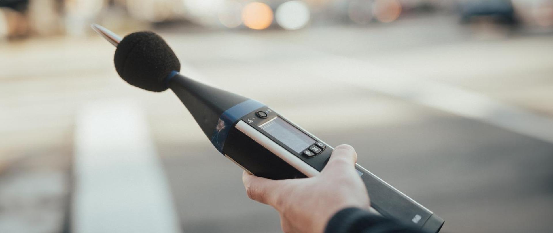 Na zdjęciu widać jak ktoś trzyma urządzenie do pomiaru hałasu. 