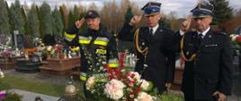 Widok z przodu. Na cmentarzu przed grobem stoją dwaj strażacy w mundurach wyjściowych i jeden strażak w ubraniu dowódczo-sztabowym. Wszyscy strażacy salutują.