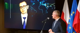 Minister cyfryzacji, Marek Zagórski, podczas konferencji prasowej przemawia do mikrofonu. W tle prezentacja multimedialna oraz flagi Polski i UE.