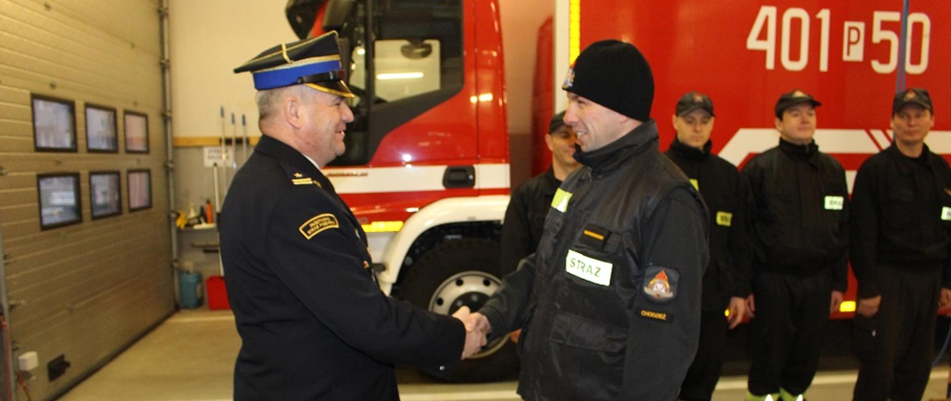 Zdjęcie przedstawia strażaków podczas uroczystej zmiany służy i wręczenia decyzji.
W tle wnętrze garażu JRG.
