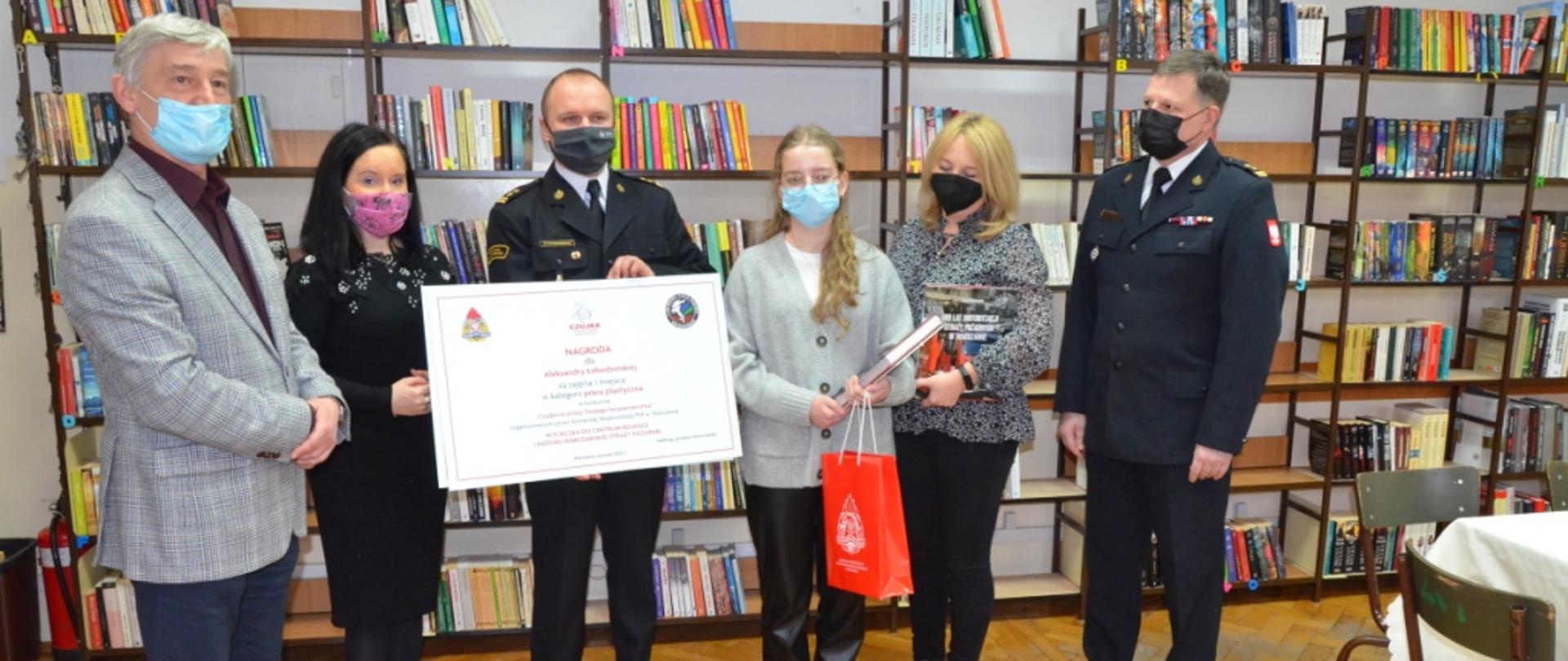 Zdjęcie wykonane w pomieszczeniu sali dydaktycznej jednej ze szkół. Na zdjęciu od lewej widać dyrektora szkoły, przedstawicielkę Komendy Wojewódzkiej Państwowej Straży Pożarnej w Warszawie, Komendanta Miejskiego Państwowej Straży Pożarnej w Radomiu, uczennicę - laureatkę konkursu, jej opiekunkę – nauczycielkę, funkcjonariusza Komendy Wojewódzkiej Państwowej Straży Pożarnej w Warszawie. Komendant Miejski Państwowej Straży Pożarnej w Radomiu trzyma przed sobą dyplom - nagrodę za zajęcie pierwszego miejsca - wycieczka do Centrum Edukacji i Historii Warszawskiej Straży Pożarnej oraz zwiedzanie Zamku Królewskiego w Warszawie. Finalistka konkursu w rękach trzyma pozostałe nagrody w postaci pamiątkowych upominków. 