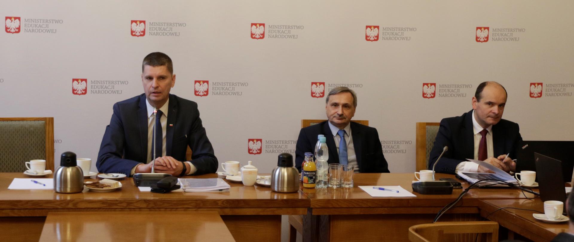 Minister edukacji i wiceminister Maciej Kopeć siedzą przy stole prezydialnym. W tle tablica z nazwą Ministerstw O edukacji Narodowej. 