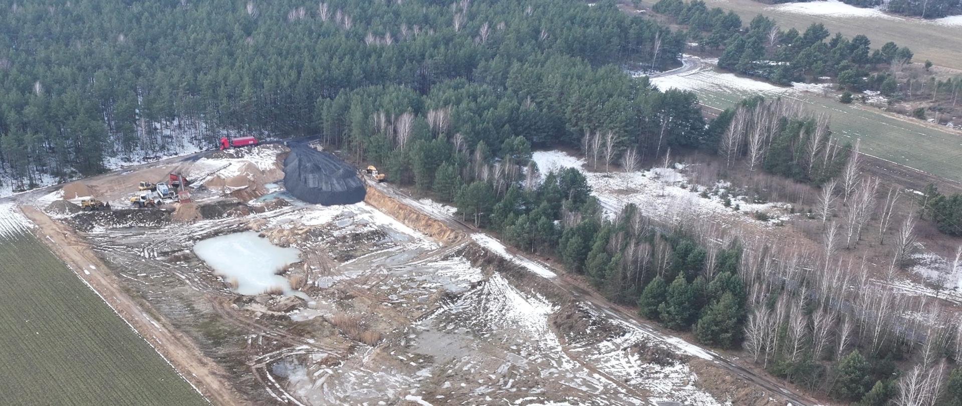 Widok wyrobiska górniczego z drona z ciężarówkami i spychaczami w czasie pracy oraz niezidentyfikowany czarny materiał deponowany w wyrobisku.