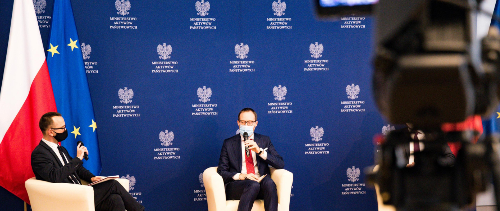 Od lewej: Karol Manys rzecznik MAP i Marek Dietl prezes GPW siedzą w fotelach podczas panelu. W tle ścianka ministerstwa. Za rzecznikiem flagi: polska i unijna. Na pierwszym planie po prawej stronie kamera w nieostrości