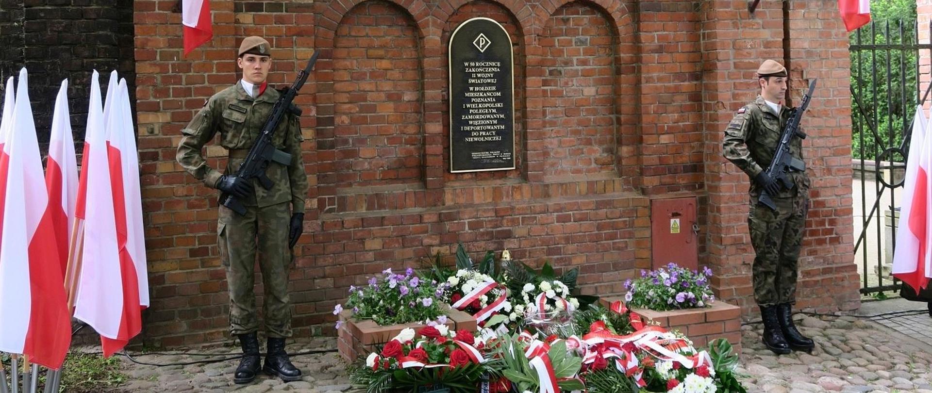 Tablica w hołdzie mieszkańcom Poznania i Wielkopolski, wisząca na murze z czerwonej cegły. pod murem złożone liczne kwiaty z biało-czerwonymi szarfami. Po bokach stojąca warta honorowa w mundurach.