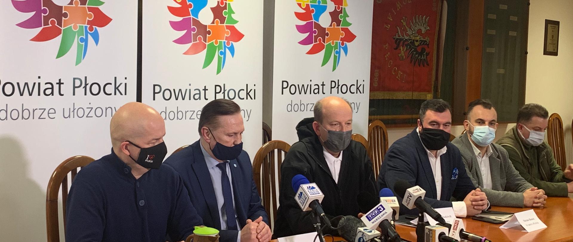 6 osób siedzących przy stole, na stole znajdują się mikrofony stacji telewizyjnych i radiowych, w tle banery z napisem Powiat Płocki