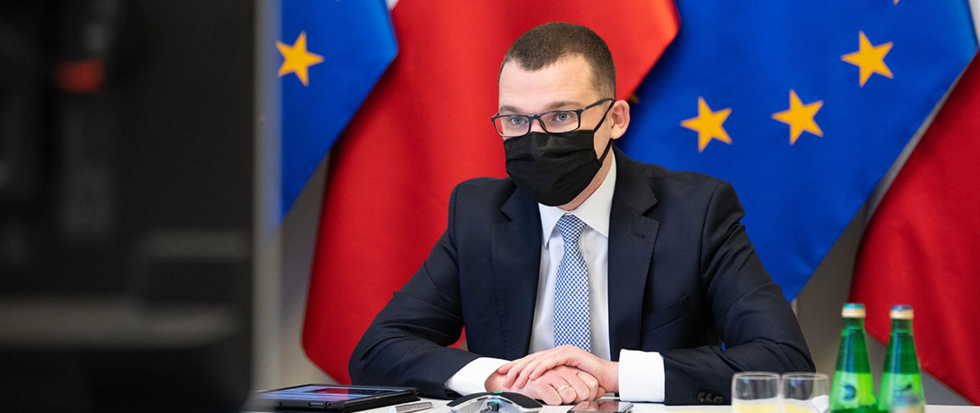 Wiceminister Paweł Szefernaker podczas posiedzenia Komisji Wspólnej Rządu i Samorządu Terytorialnego w formie wideokonferencji na tle flag Polski i Unii Europejskiej.