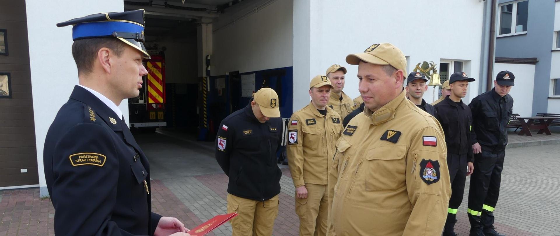 Zdjęcie przedstawia st. kpt. Roberta Czarneckiego p.o. Komendanta Powiatowego PSP w Brodnicy wręczającego st. ogn. Przemysławowi Krajnikowi list gratulacyjny wraz informacją o przyznaniu nagrody z okazji jubileuszu pracy zawodowej. W tle strażacy podziału bojowego. Za nimi budynek JRG Brodnica.