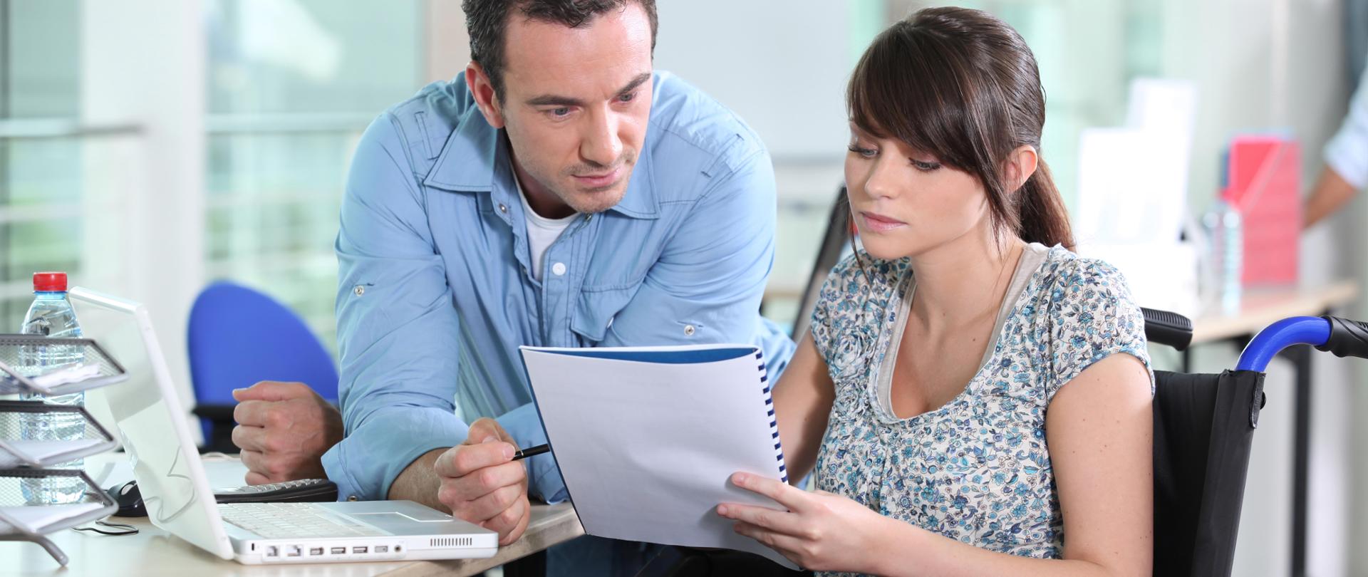 Kobieta na wózku inwalidzkim trzyma zeszyt i rozmawia z mężczyzną który opiera się o biurko na którym jest laptop