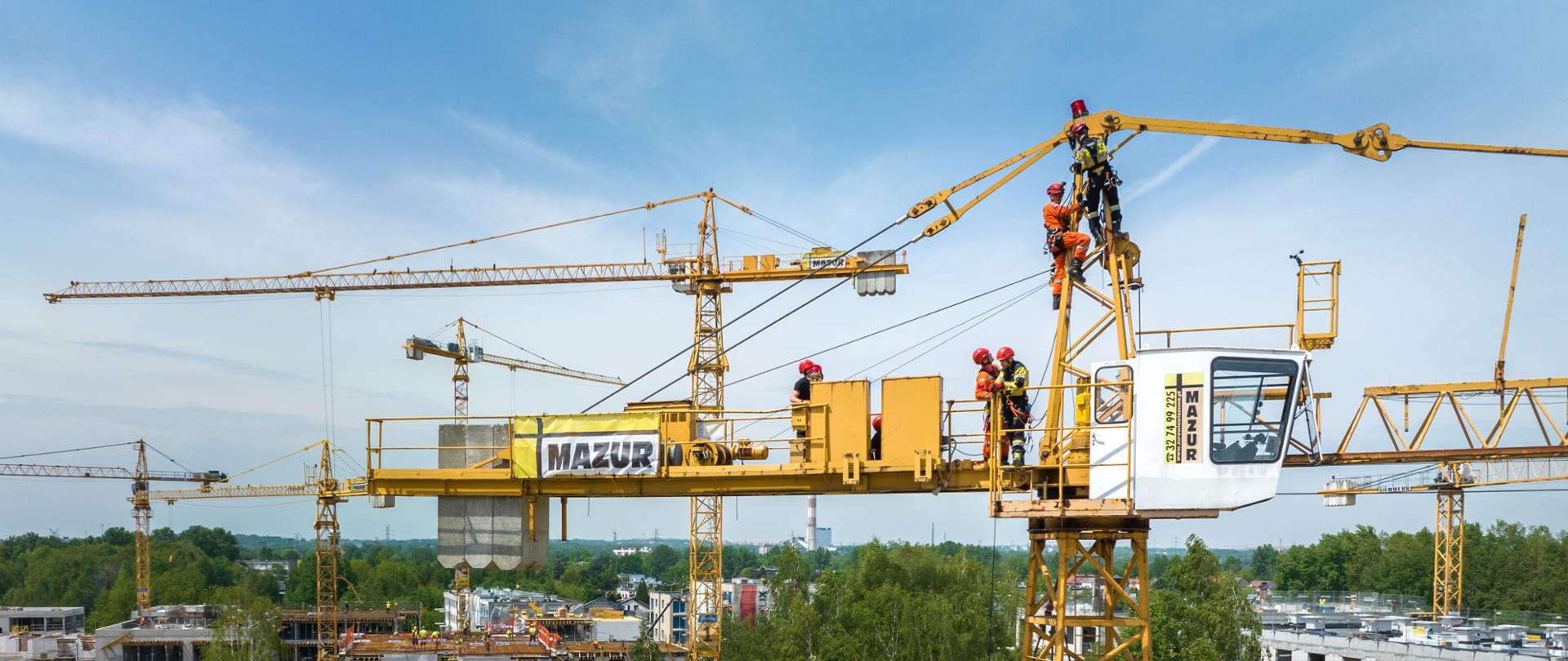 Strażacy ze specjalistycznych grup ratownictwa wyskościowego ćwiczą na żurawiu ustawionym na placu budowy. W tle budowane bloki i drzewa.