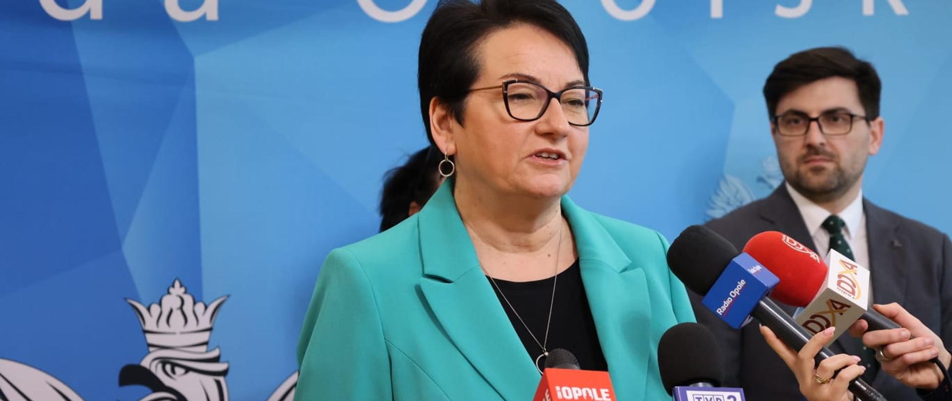 Wojewoda Opolski Monika Jurek w trakcie konferencji dotyczącej środków na realizację zadań w zakresie wychowania przedszkolnego dla Województwa Opolskiego na 2024 rok. Wojewoda mówi do mikrofonów, które widać z prawej strony fotografii. 