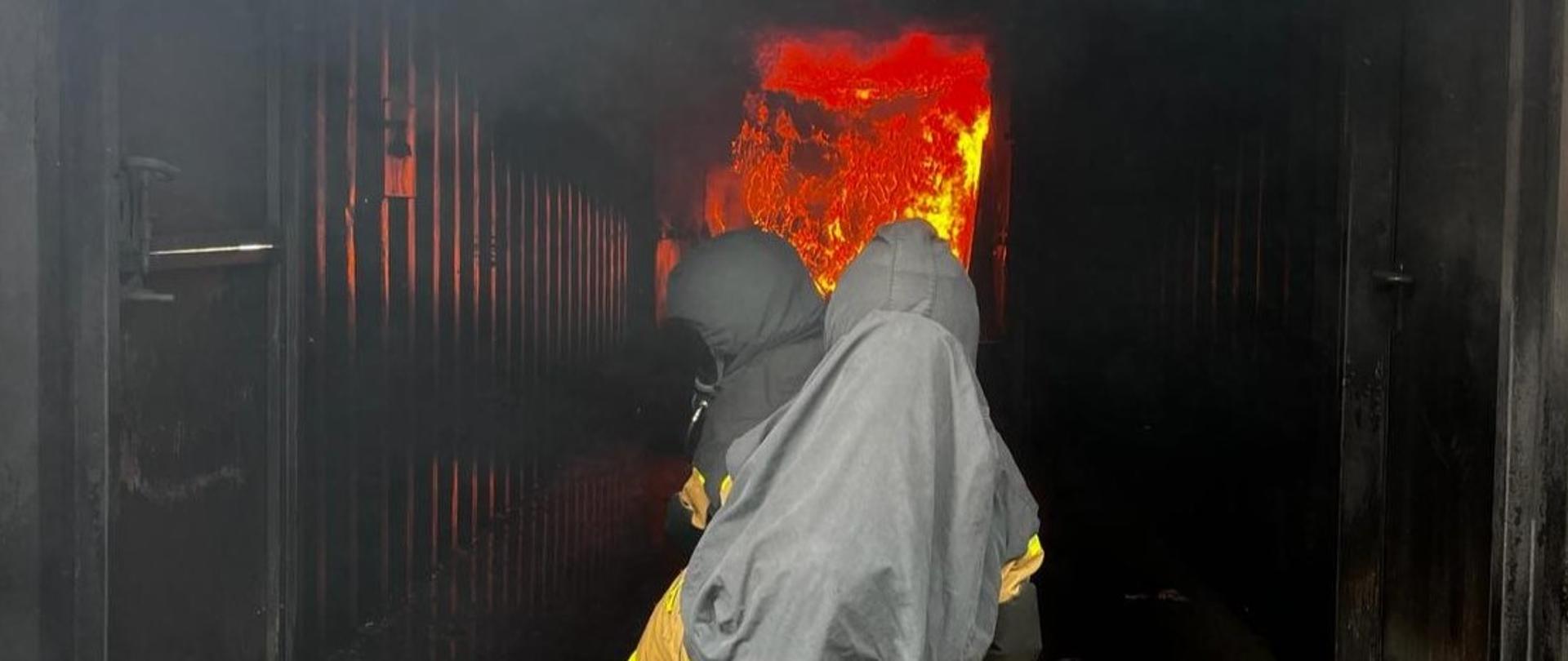 Strażacy ochotnicy w ubraniu bojowym w hełmach na głowach ćwiczą operowanie prądami wody w komorze rozgorzeniowej podczas ćwiczeń w pożarach wewnętrznych. Ćwiczenia przeprowadzone na poligonie ratowniczym Komendy Wojewódzkiej Państwowej Straży Pożarnej w Opolu.