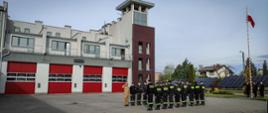 Na tle budynku strażnicy, w środkowej części w dwóch rzędach stoją strażacy w umundurowaniu koszarowym. Po prawej stronie flaga państwowa wciągnięta na maszt.