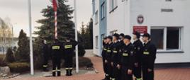 Strażacy stoją na baczność w dwóch rzędach oddając honor podczas wciągania flagi Polski na maszt przez poczet flagowy.
