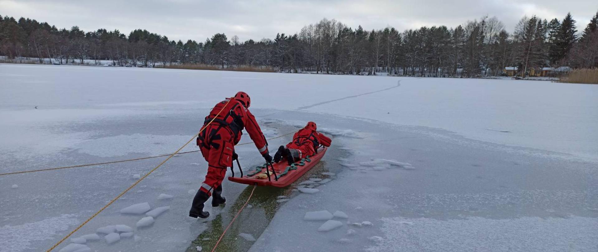 Strażacy w suchych skafandrach ratowniczych ćwiczą elementy ratownictwa na lodzie z wykorzystaniem sań wodno- lodowych. Strażacy są asekurowani z brzegu linami.