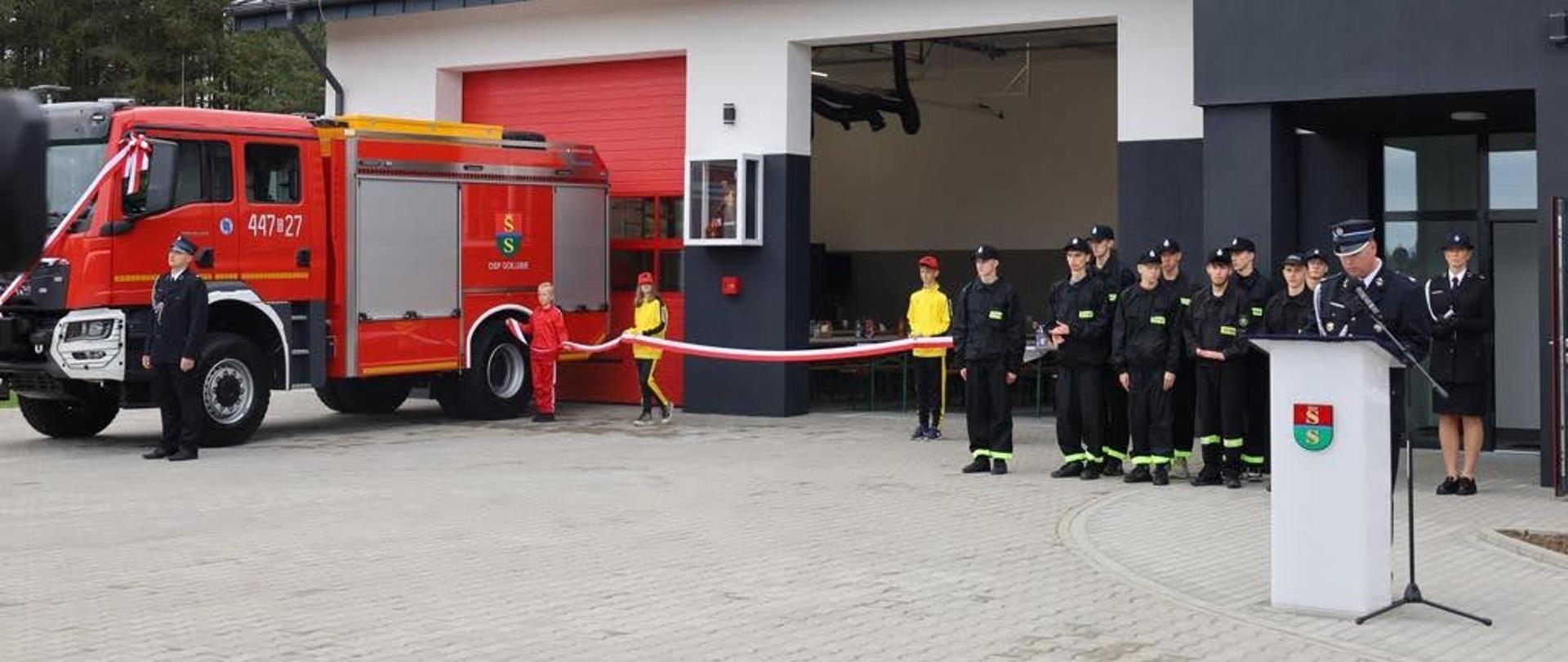 Samochód gaśniczy stoi przed remizą ochotniczej straży pożarnej w Gołubiu obok drzwi auta stoi strażak za nim stoi dzieci w stroju koloru czerwonego oraz w żółto - czarne dresy następnie w dwóch rzędach stoją młodzi strażacy przy mównicy przemawia strażak ochotniczej straży pożarnej. 