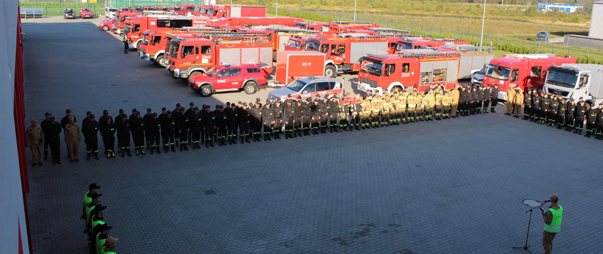 Zdjęcie przedstawia zbiórkę przed ćwiczeniami strażaków z województwa podkarpackiego biorących udział w ćwiczeniach. W tle widoczne są samochody Straży Pożarnej.