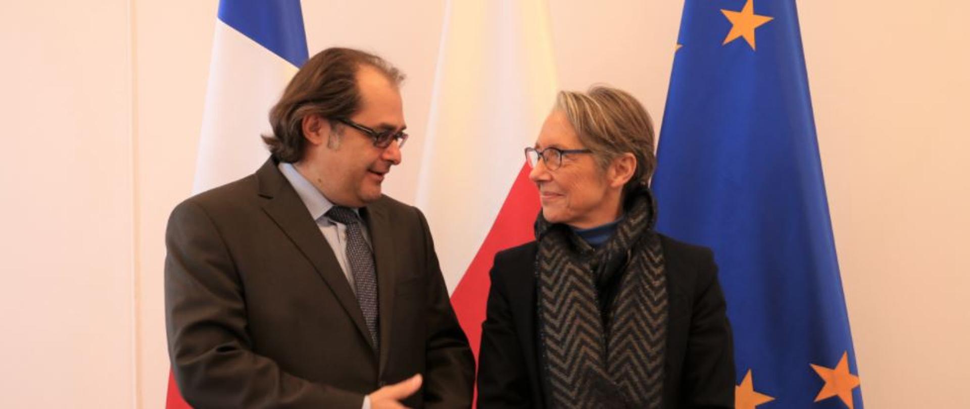Na zdjęciu od lewej strony znajduje się minister Marek Gróbarczyk oraz minister Élisabeth Borne. Ministrowie rozmawiają na tle trzech flag: Francji, Polski i Unii Europejskiej.