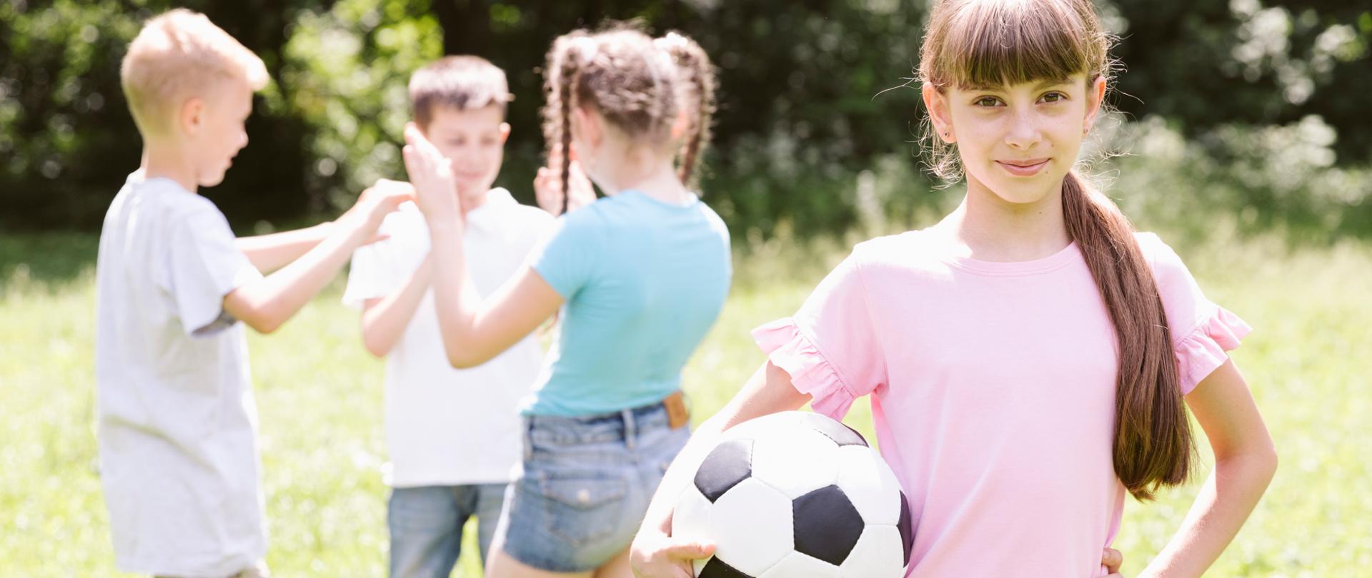 Dziewczynka trzymająca piłkę do nogi, na drugim planie troje innych dzieci rozmawiających ze sobą.