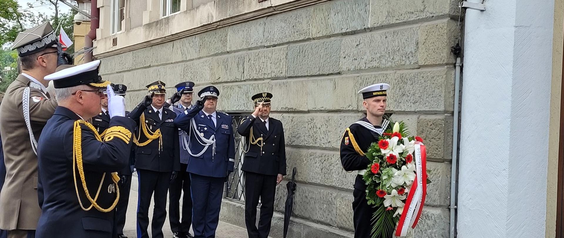 Przedstawiciele służb mundurowych oraz wojska oddają honory przed nimi stoi żołnierz z wiązanką.