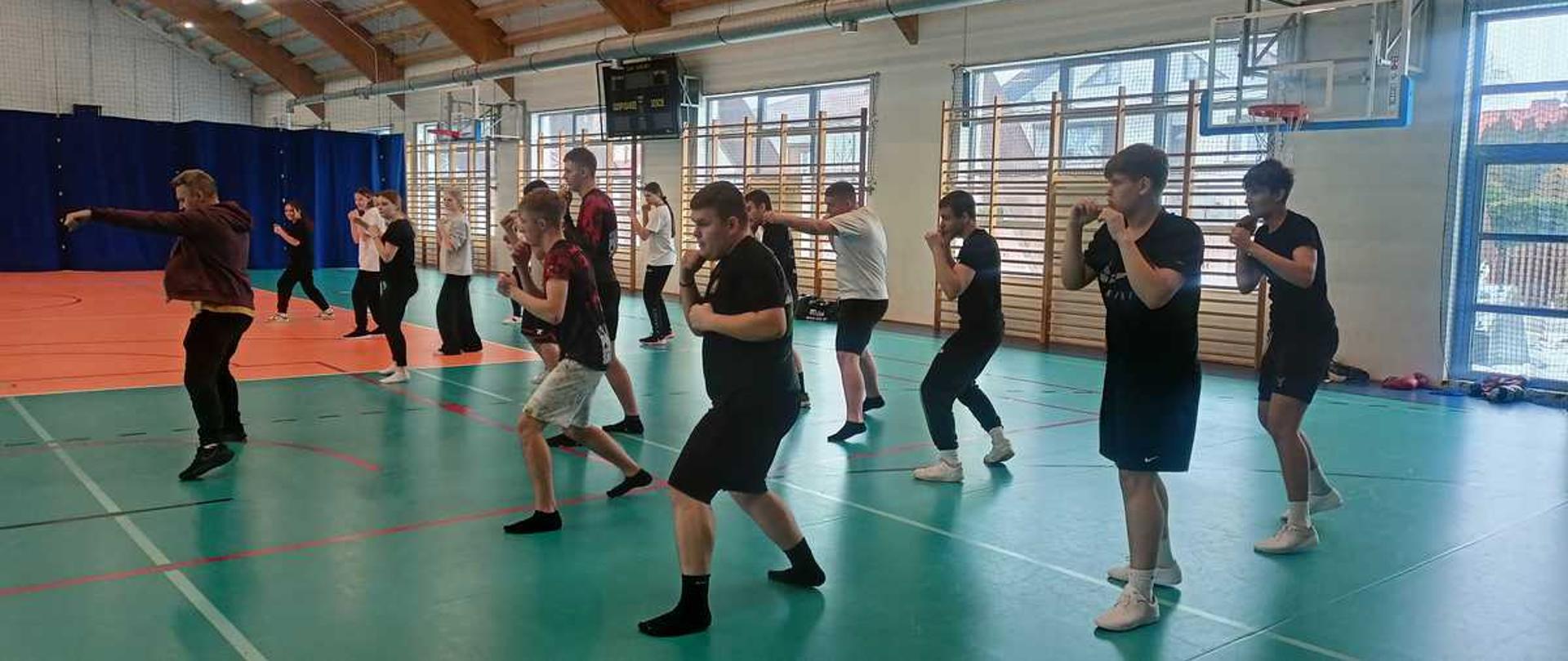 Zdjęcie przedstawia zajęcia sportowe w dużej hali sportowej. Grupa młodych ludzi ustawionych w kliku rzędach wykonuje ćwiczenia przypominające trening bokserski. Wszyscy stoją w lekkim rozkroku z uniesionymi rękoma, wykonując ruchy ciosów w powietrzu. Na pierwszym planie widać instruktora, który prowadzi trening i pokazuje ruchy.