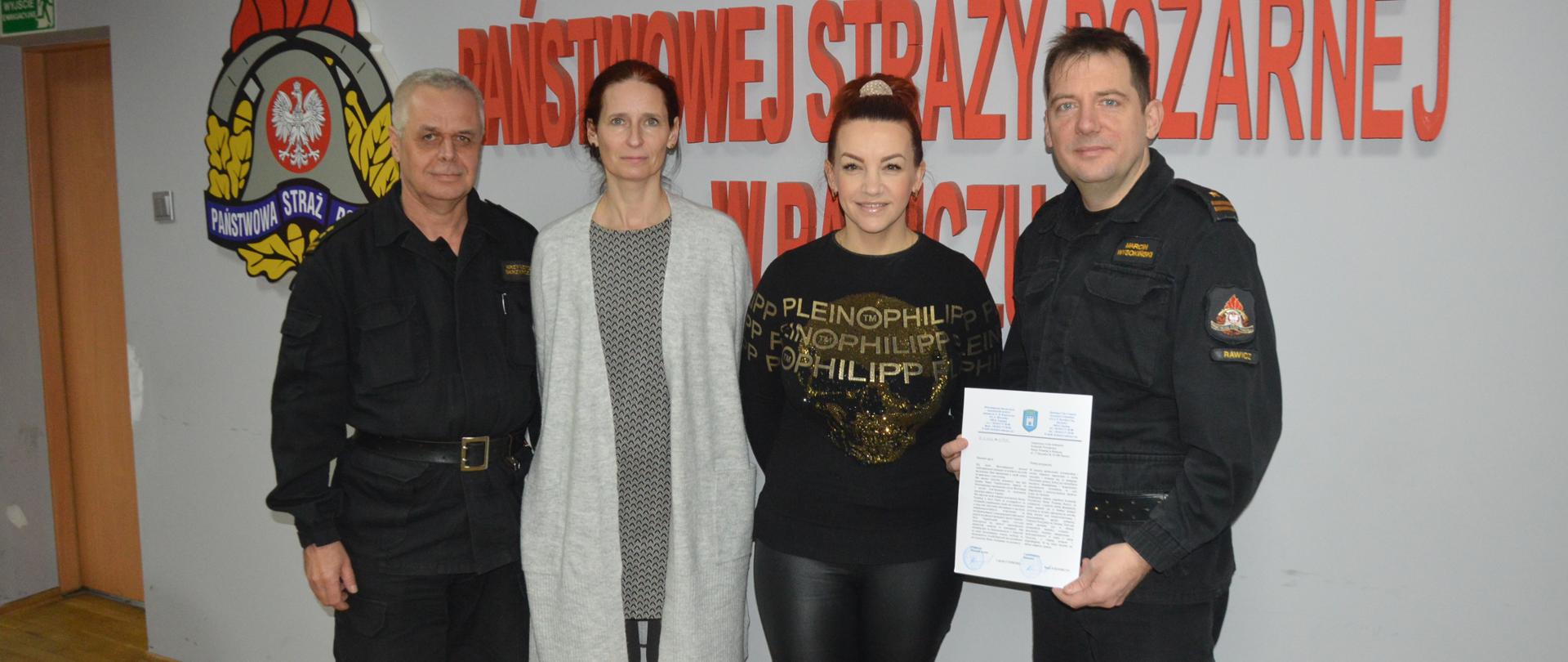 List od burmistrza Żytomierza. Świetlica Komendy Powiatowej PSP w Rawiczu. Na tle ściany, na której znajduje się czerwony napis "KOMENDA POWIATOWA PAŃSTWOWEJ STRAŻY POŻARNEJ W RAWICZU" oraz logo PSP, stoją obok siebie 4 osoby - 2 strażaków w ubraniach dowódczo-sztabowych oraz dwie kobiety w strojach cywilnych. Strażak stojący z prawej strony trzyma w rękach list. 