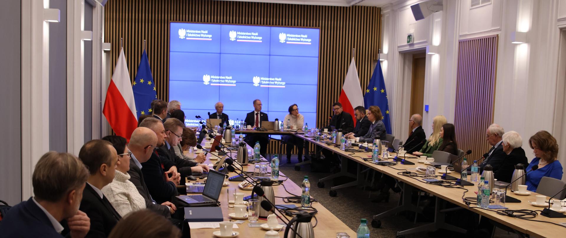 Widok z tyłu na salę, przy ustawionych w czworokąt stołach siedzą ludzie, przed nimi małe mikrofony na stojakach, z przodu sali pod wbudowanym w ścianę wielkim ekranem siedzi wiceminister Szeptycki.