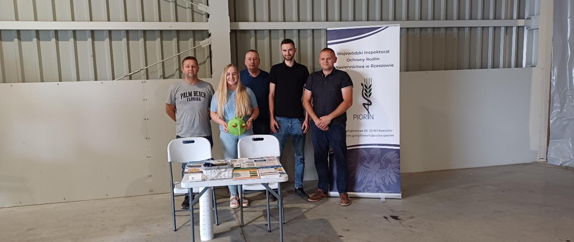 Grupa ludzi stojąca w szarym pomieszczeniu. Czterech mężczyzn i jedna kobieta która trzyma w rękach zieloną maskotkę. Za nimi stoi biało-granatowy baner z logiem instytucji. Przed nimi jest stolik a na nim ulotki