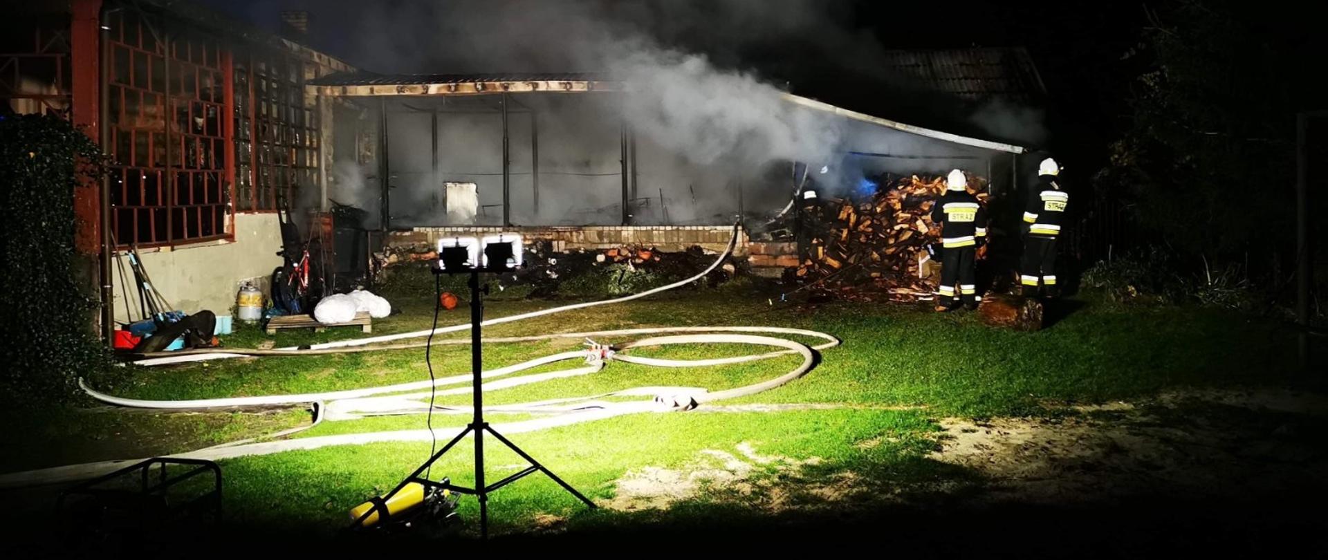 Teren akcji gaśniczej, oświetlony stojącą na przenośnym stojaku lampą. W świetle lampy widoczny niski budynek, z którego wydobywa się dym, do środka budynku wprowadzony jest wąż gaśniczy. Budynek jest niski i jest częścią większego kompleksu budynków. Przed budynkiem stoi dwóch strażaków w białych hełmach.