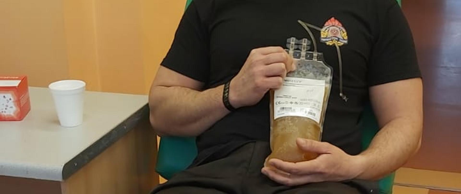 Strażak w ubraniu koszarowym siedzący na fotelu w centrum krwiodawstwa. Na twarzy ma maseczkę, a w ręku trzyma pobrane osocze.