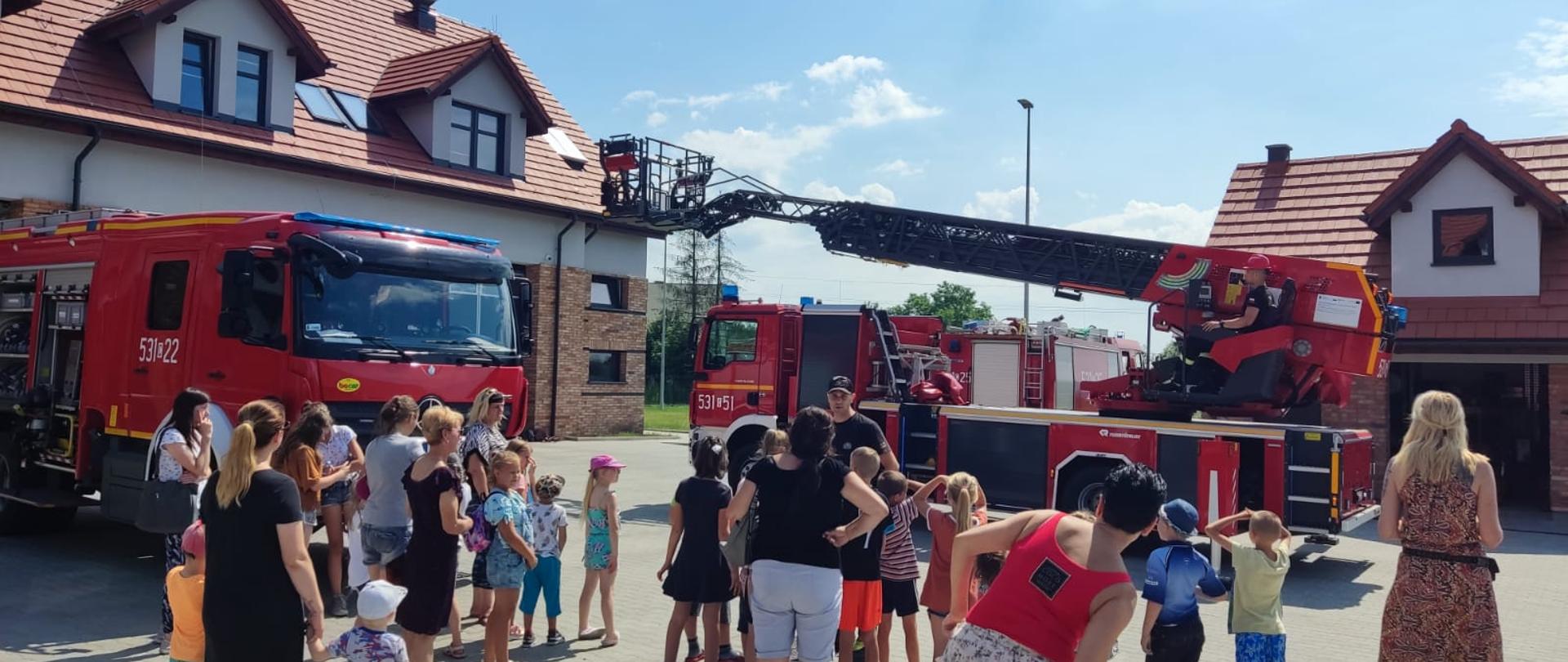 Na placu Komendy stoi grupa dzieci z opiekunami, strażak opowiada im o drabinie mechaniczne, która rozkłada się w tle.
