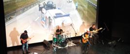 Koncert „Kapeli PSP” trzech gitarzystów oraz perkusista na scenie. W tle prezentacja na której widoczna jest karetka.