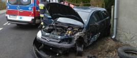Zdjęcie przedstawia czarny samochód osobowy marki opel z rozbitym przodem i podniesioną maską. Obok auta stoi karetka oraz strażak i ratownik medyczny.