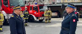 Zdjęcie zrobione na placu komendy przedstawia Komendanta Wojewódzkiego, któremu melduje dowódca uroczystości w tle samochody ratowniczo-gaśnicze oraz strażacy stojący na baczność.