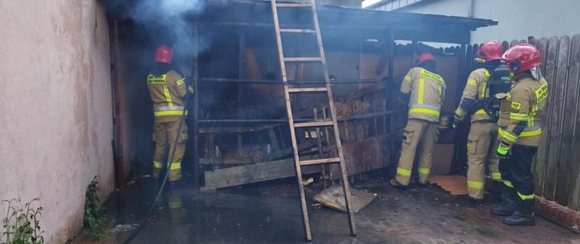 Zdjęcie wykonane w dzień w trakcie działań gaśniczych przy palącym się drewnie pod wiatą. Przy wiacie stoi dwóch strażaków w piaskowych mundurach gaszących drewno. Wiata jest nie duża wypełniona po brzegi drewnem.