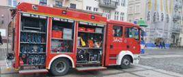 Na zdjęciu widać samochód gaśniczy należący do JRG 1 Kalisz. Samochód ma kolor czerwony, ma oznakowanie oraz lampy ostrzegawcze kolory niebieskiego. Skrytki zabudowy są otwarte. Pojazd stoi na płycie rynku. W tle widać zabudowania Głównego Rynku.