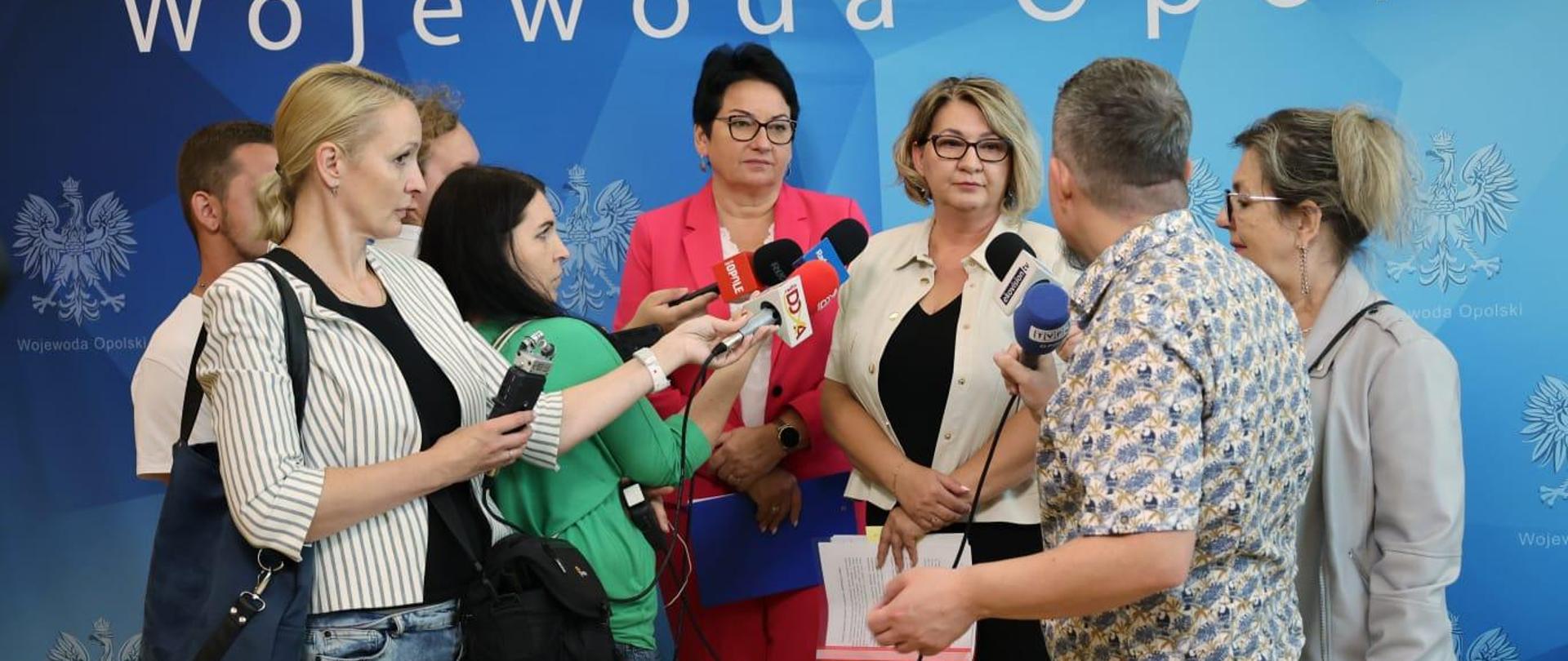 Wojewoda Opolski Monika Jurek oraz Dyrektor Wydziału Polityki Społecznej Ewa Pawlinów w trakcie konferencji prasowej. Po obu stronach stoją dziennikarze, którzy trzymają mikrofony skierowane w stronę Dyrektor Ewy Pawlinów. W momencie robienia zdjęcia, jeden z dziennikarzy zadawał pytanie. 