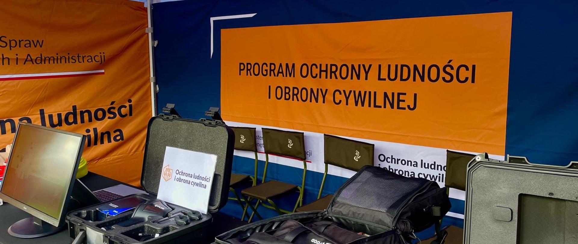 Stoisko edukacyjne programu ochrony ludności i obrony cywilnej. Na stołach znajdują się walizki ze sprzętem, monitor, plecaki i inne elementy wyposażenia prezentowane uczestnikom wydarzenia. W tle widoczny jest baner z napisem „Program Ochrony Ludności i Obrony Cywilnej”.