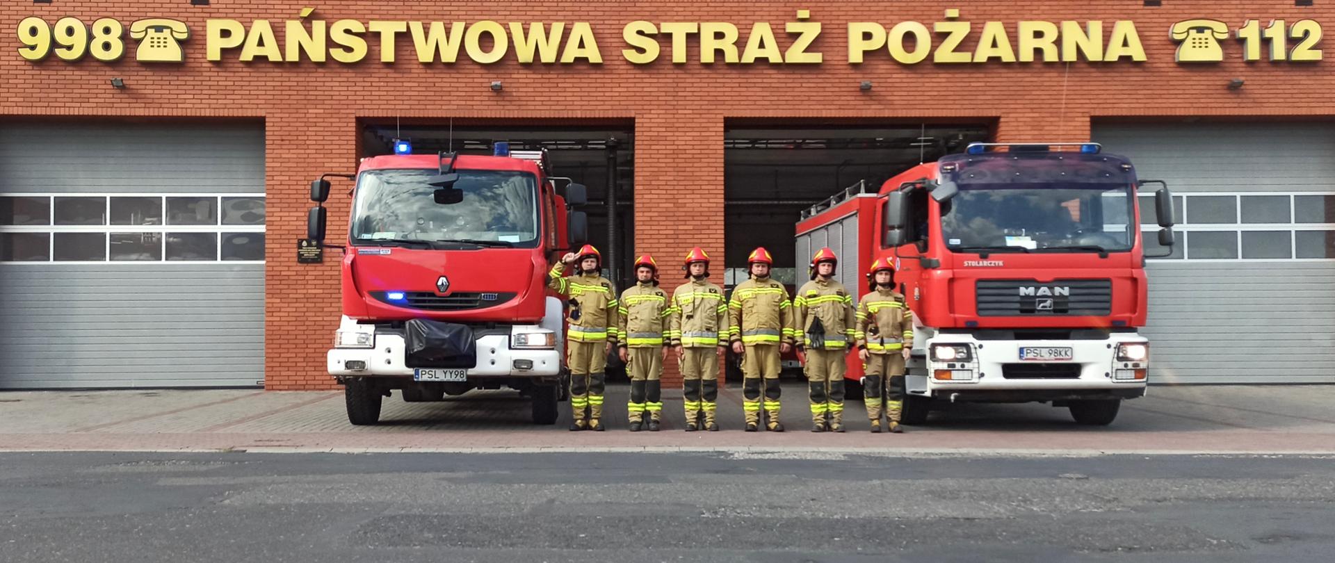 Sześciu strażaków w żółtych ubraniach specjalnych z czerwonymi hełmami na głowie stoi w szeregu pomiędzy dwoma samochodami pożarniczymi koloru czerwonego z włączonymi światłami błyskowymi. W tle budynek Komendy Powiatowej Państwowej Straży Pożarnej w Słupcy z czerwonej cegły klinkierowej z napisem "998 Państwowa Straż Pożarna 112"