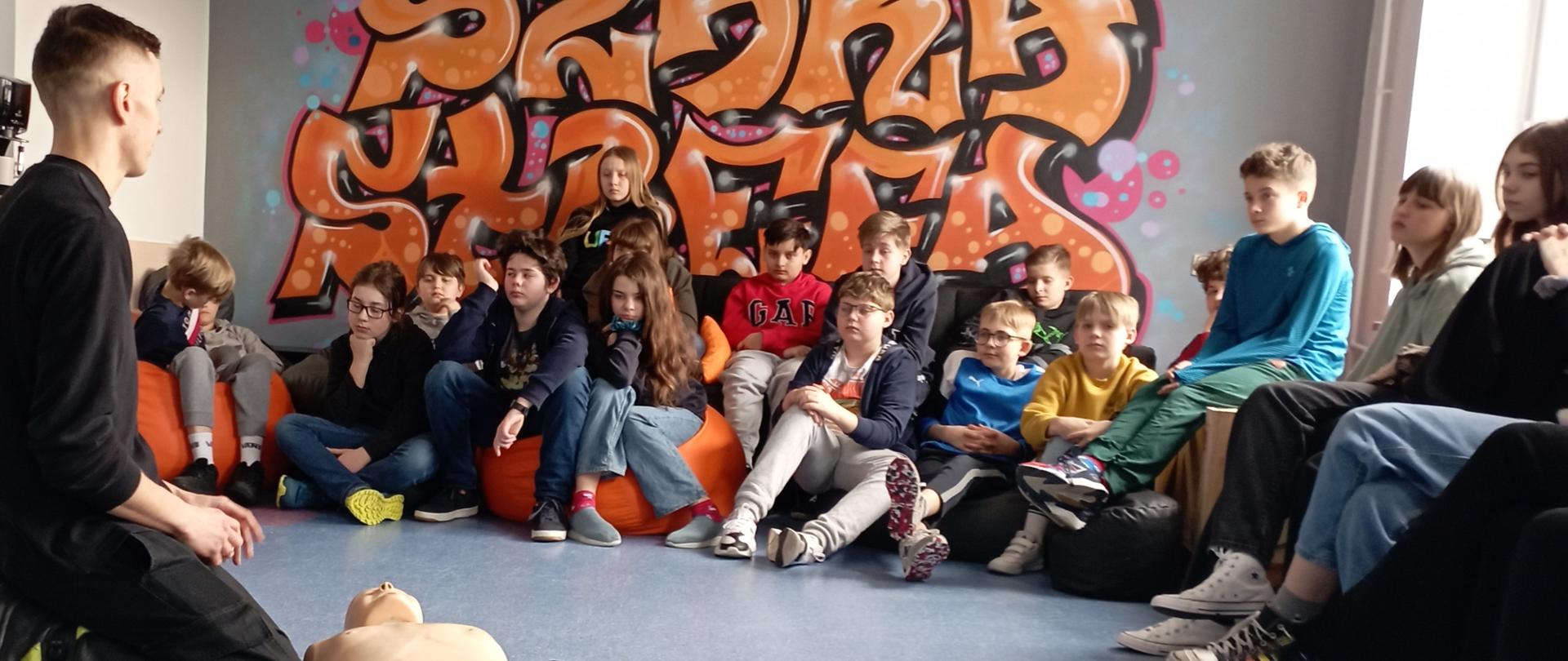 Na zdjęciu przedstawiono warsztaty z uczniami w szkole podstawowej w ramach profilaktyki bezpieczeństwa. Na pierwszym planie ratownik medyczny prezentuje prawidłowy przebieg resuscytacji krążeniowej, w tle młodzież szkolna.