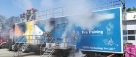 Kontener z którego szczelin wydobywa się dym, na jego dachu ogrodzonym barierkami stoi trzech strażaków w chełmach. Na ścianach kontenera namalowane postacie strażaków oraz napis: Fire Training