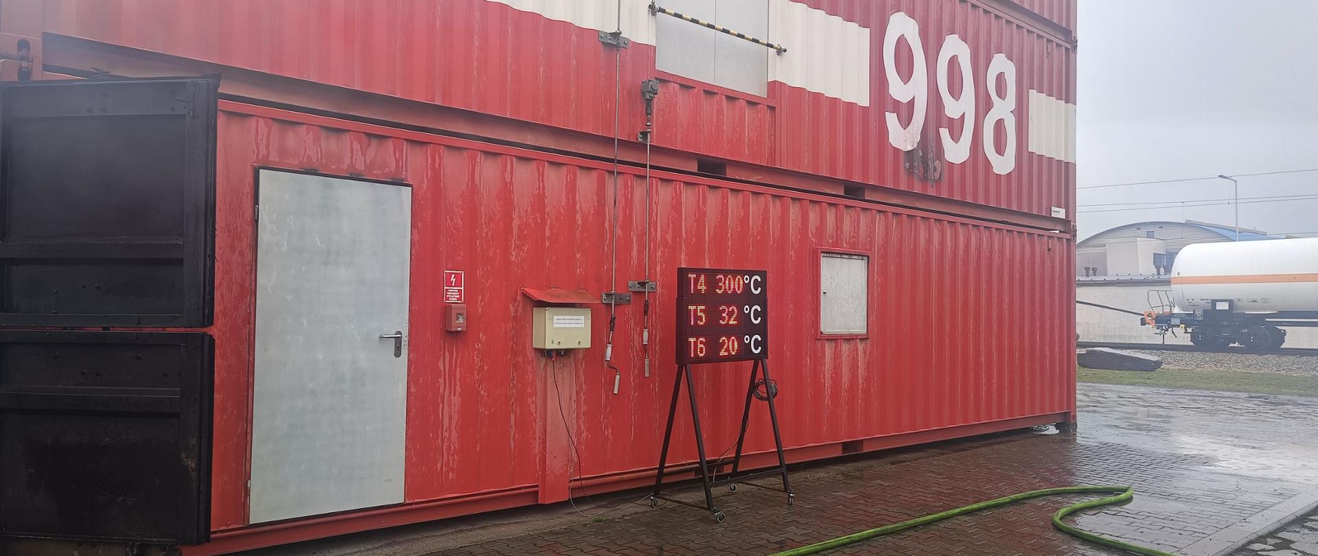 Widok na kontenerową komorę rozgorzeniową przed którą ustawiona jest elektroniczna tablica informująca o temperaturze panującej w różnych lokalizacjach