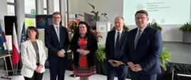 Pięć osób stoi obok siebie w formalnym otoczeniu. Od lewej: kobieta w jasnej marynarce, dwóch mężczyzn w garniturach i krawatach, kobieta w czerwonej sukience z wzorami i ciemnej narzutce oraz kolejny mężczyzna w garniturze. Za nimi widać ekran z napisem "Branżowe Centrum Umiejętności w Dziedzinie Ogrodnictwa Bielsko-Biała" oraz flagi Polski i Unii Europejskiej.