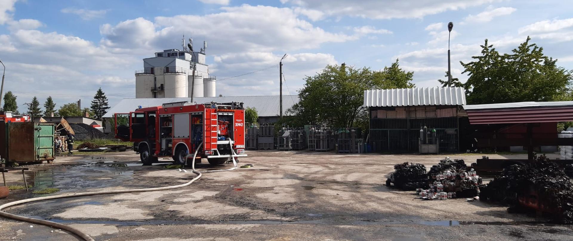 Zdjęcie przedstawia samochód ratowniczo-gaśniczy z rozwiniętą linią gaśniczą, stojący przed halą magazynową.. Plac jest mokry, z prawej strony widać pozostałości po pożarze. 