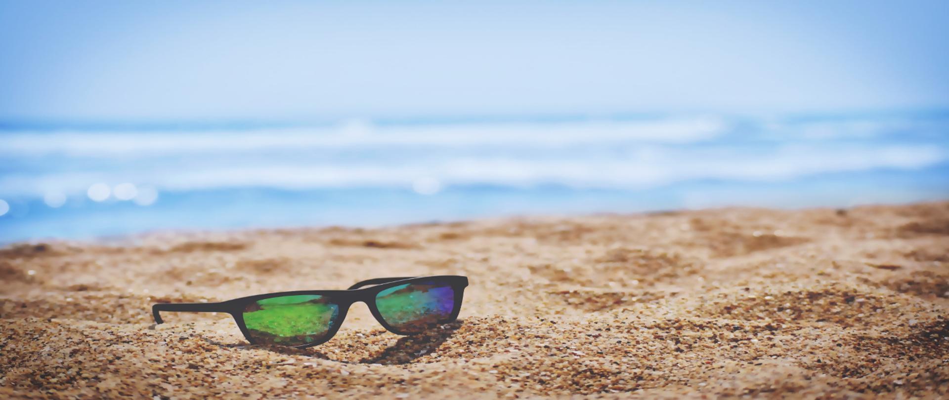 Zdjęcie przedstawiające plażę, na piasku leżą okulary przeciwsłoneczne, a w oddali widać morze.