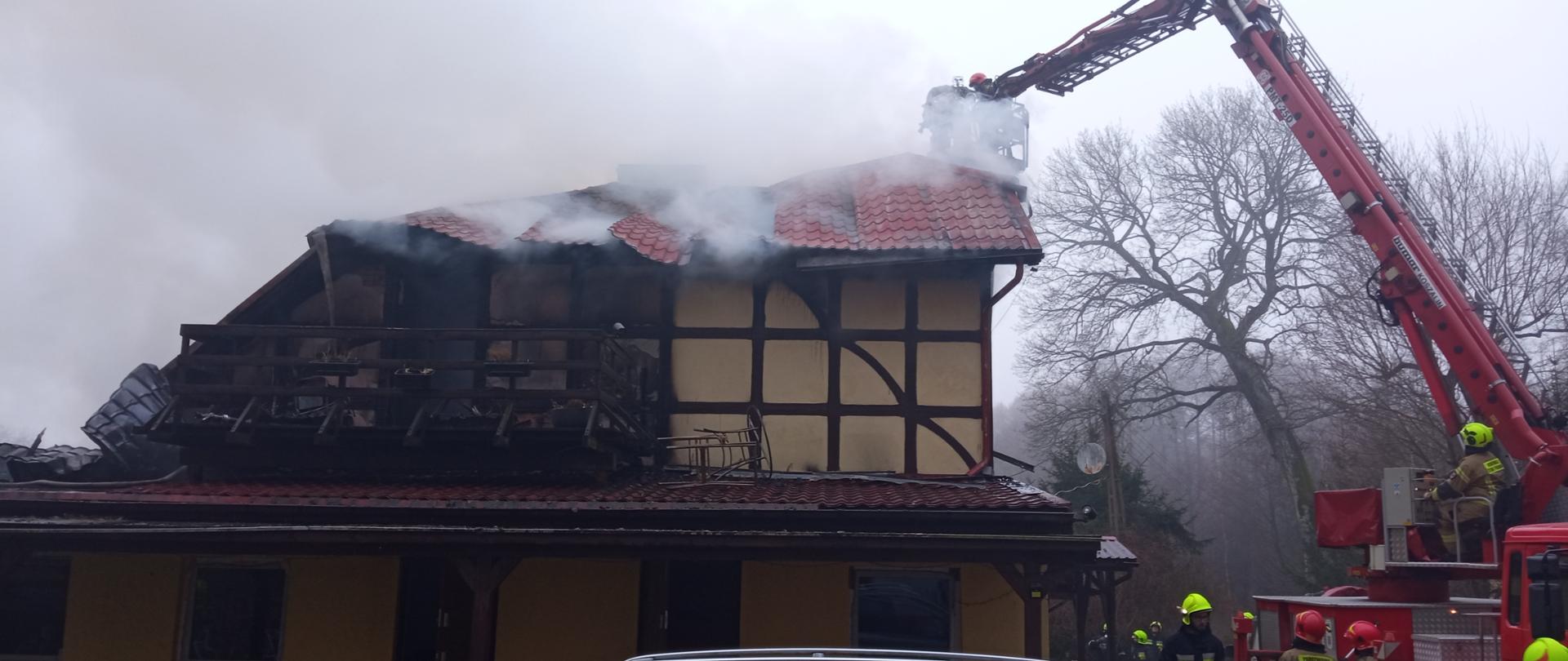 Fotografia przedstawia budynek z którego wydobywają się kłęby dymu. Konstrukcja dachu budynku częściowo zawalona, po prawej stronie kadru widać ramię rozstawionego podnośnika, w koszu podnośnika widoczny strażak.