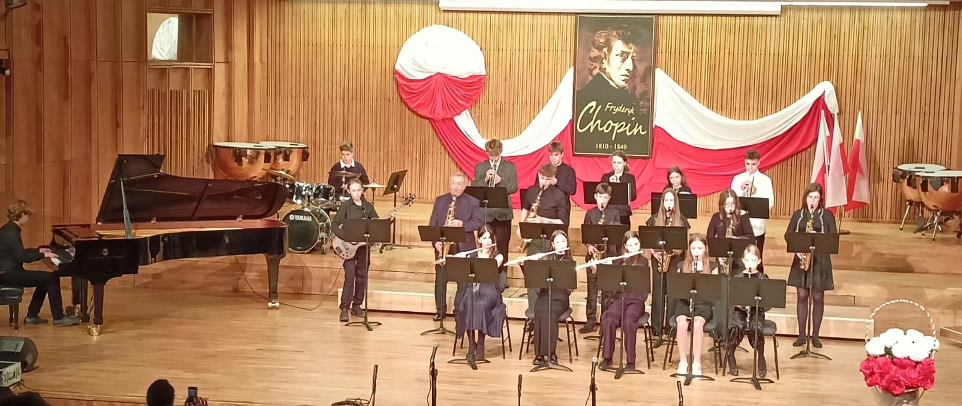 Na środku znajduje się zespół instrumentów dętych składający się z uczniów PSM I st. im. F. Chopina w Szczytnie. W tle widać obraz F. Chopina na tle polskiej flagi. Po lewej stronie znajduje się uczeń grający na fortepianie. Na dole widać publiczność.