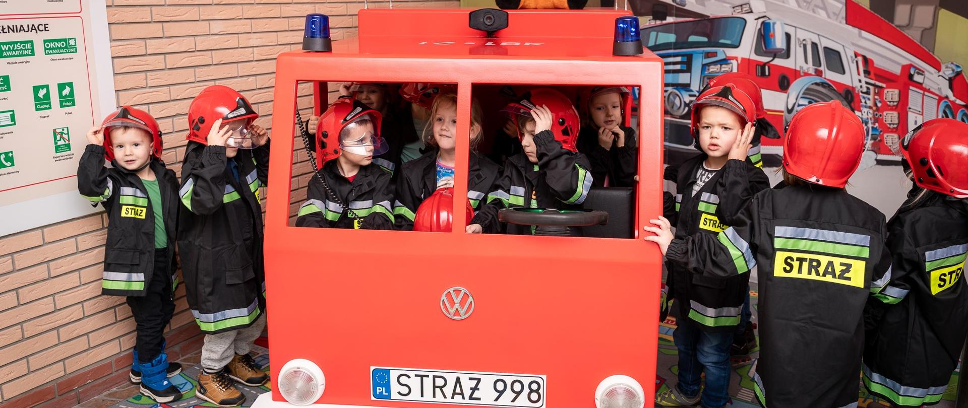 Zdjęcie wykonane we wnętrzu sali edukacyjnej "OGNIK" w KP PSP w Koluszkach. Na zdjęciu widoczne są dzieci, które bawią się we wnętrzu drewnianego samochodu pożarniczego. 