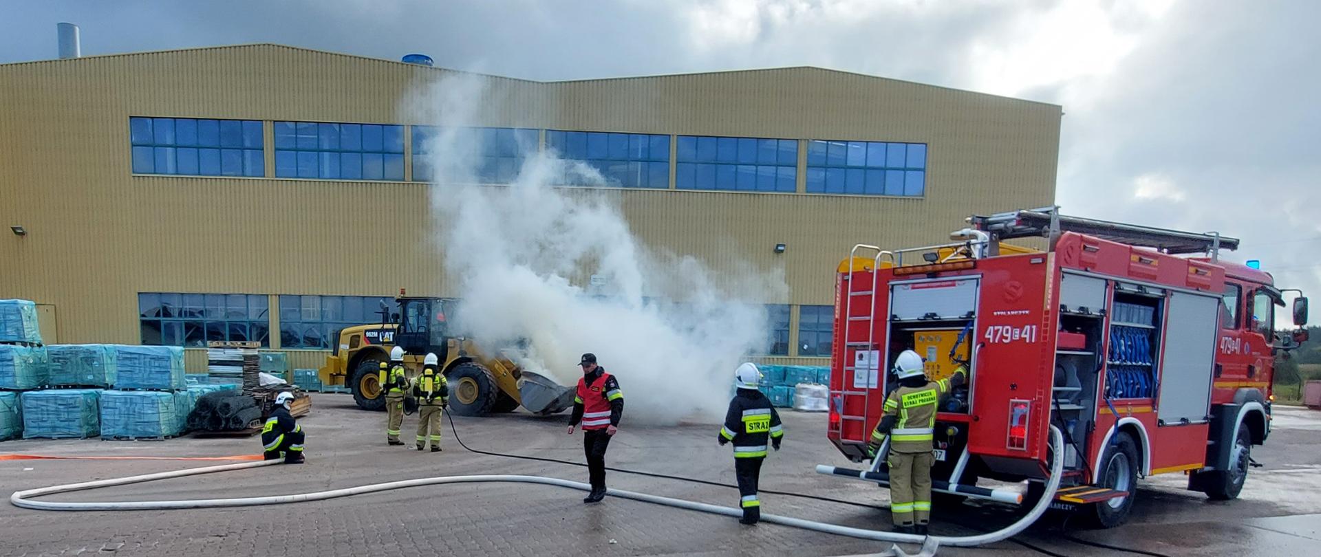 Strażacy w specjalnych ubraniach, prowadzący działania gaśnicze zadymionej ładowarki. Dwóch strażaków podaje prąd wodny, kolejnych dwóch rozwija drugą linię węży, jeden strażak obsługuje autopompę pojazdu gaśniczego w kolorze czerwonym. W polu widzenia również strażak ubrany w czerwoną kamizelkę oceniający prowadzone działania. W tle zakład produkcyjny. 
