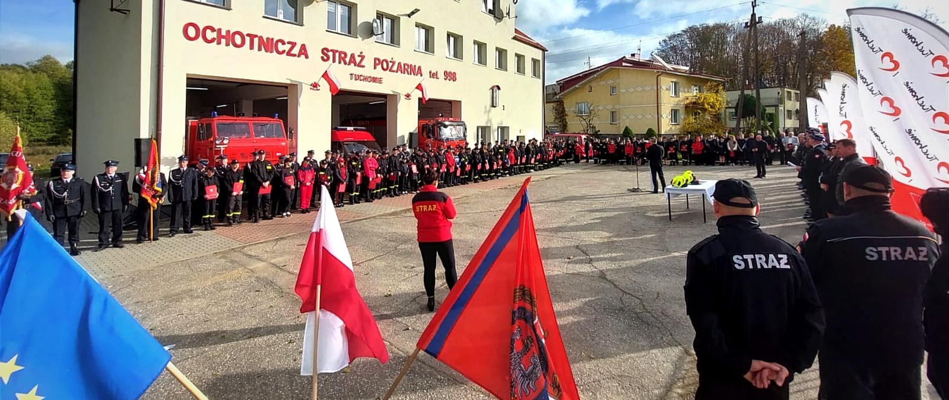 Zdjęcie przedstawia ustawionych w dwuszeregu członków OSP i MDP w umundurowaniu przed budynkiem remizy OSP w Tuchomiu. Przed nimi ustawieni są zaproszeni goście oraz mównica, przy której przemawia funkcjonariusz PSP w umundurowaniu wyjściowym
